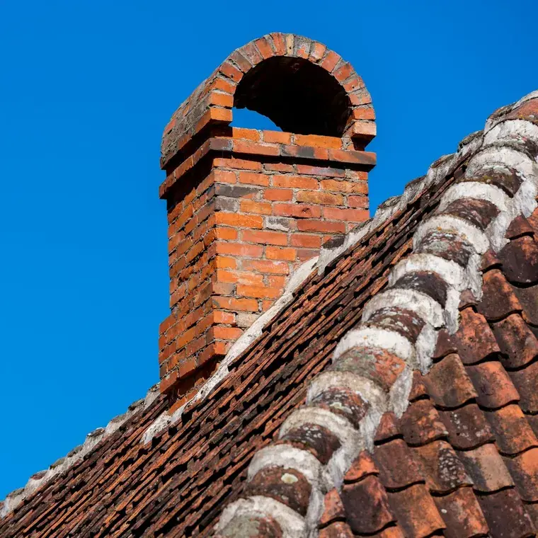 A brick chimney stack with leadwork and ridge tiles against a blue sky